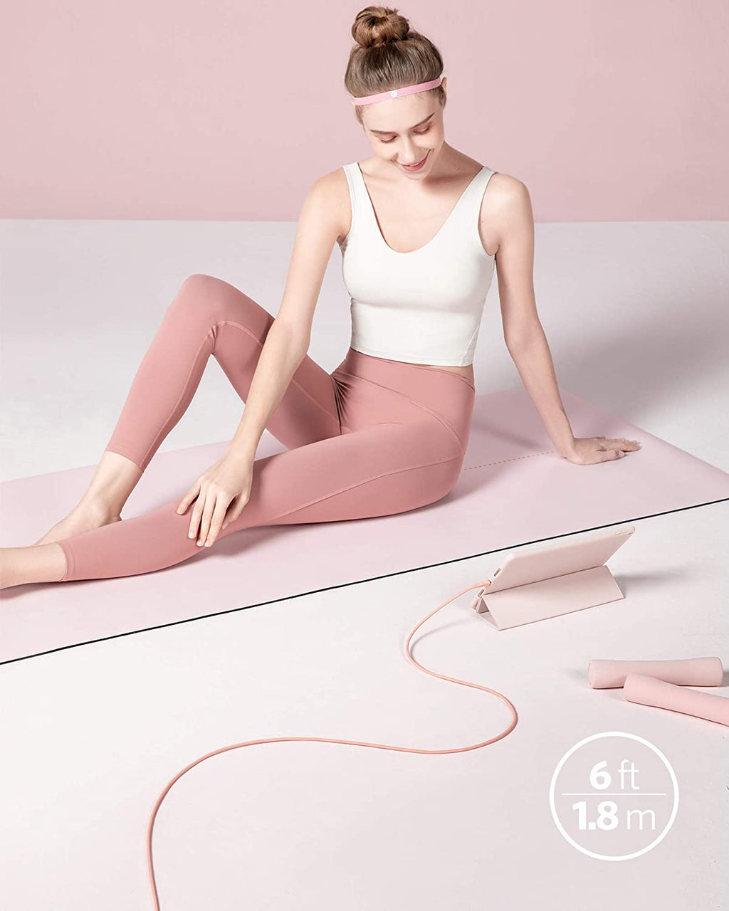 Woman in pink leggings and white tank top sitting on a pink mat with a pink yoga strap, on a light pink background.