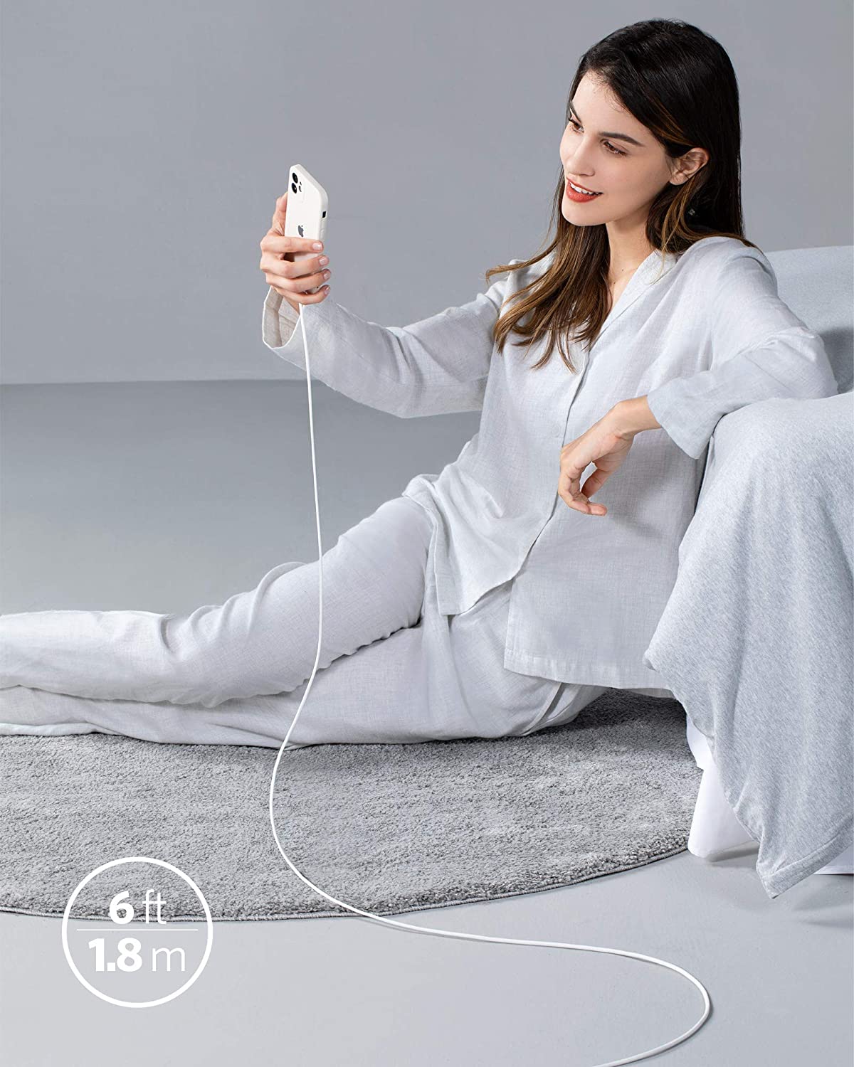 Woman sitting on a gray couch holding a phone with a long cable, wearing a white outfit.