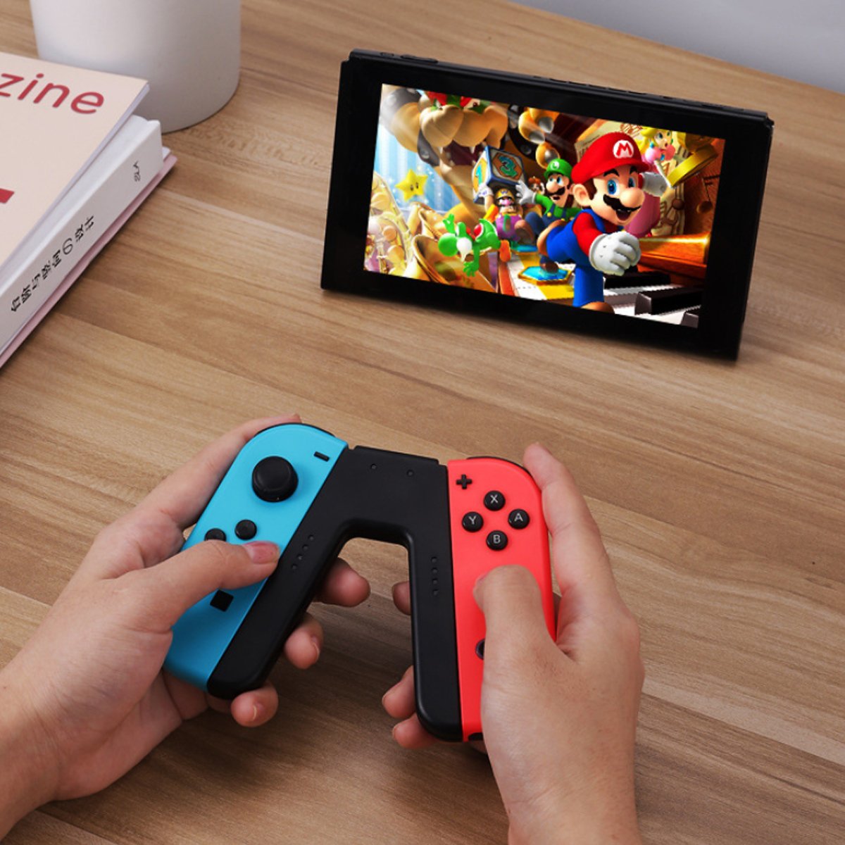 Hands holding red and blue Nintendo Switch controllers with a screen displaying a game on a wooden table.