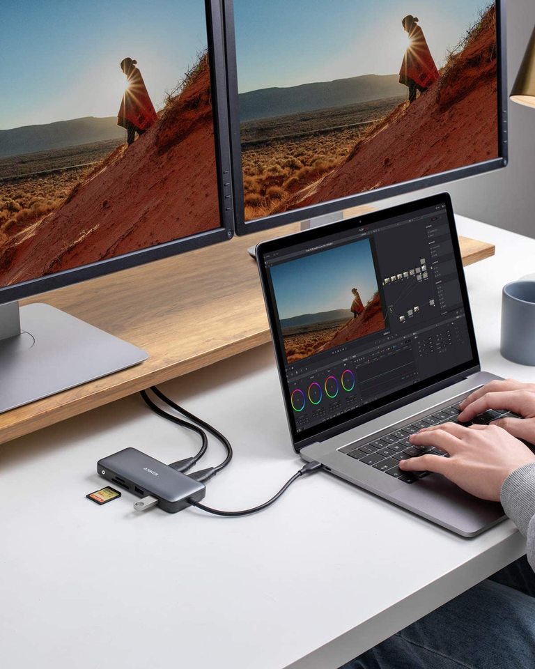 Person using a laptop with a USB hub connected to two monitors displaying scenic images.